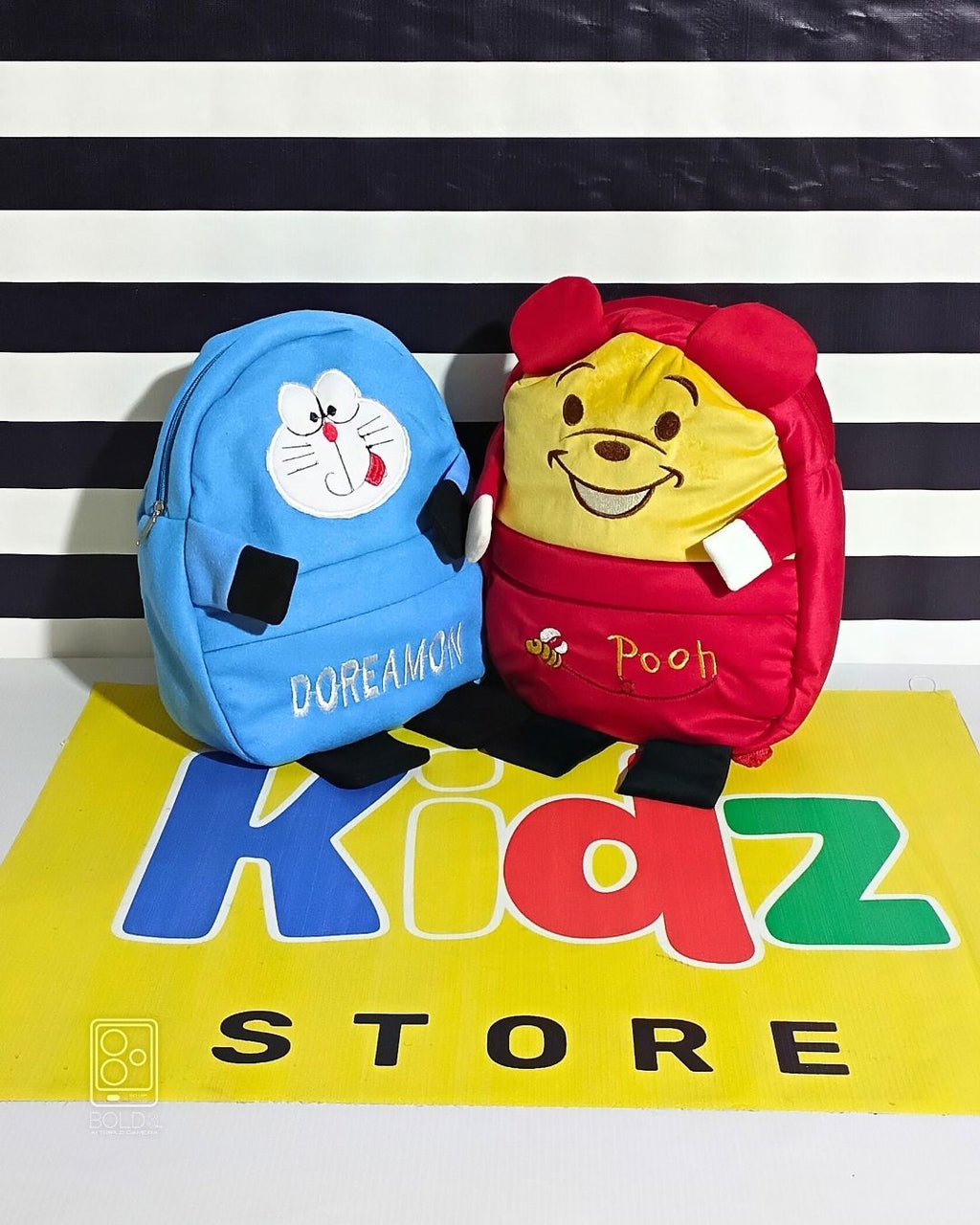 Two small backpacks, one blue with Doraemon design and one red with Pooh design, on a striped background with 'Kiaz Store' branding.