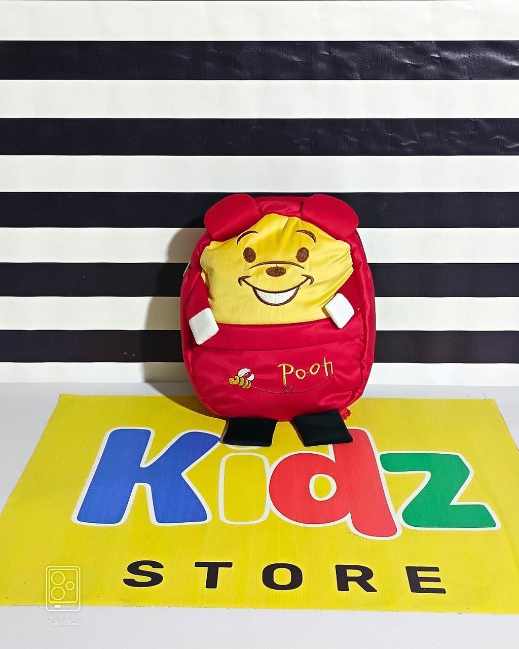 Plush toy resembling a bear character in a red outfit on a striped background with 'Kidz Store' branding.